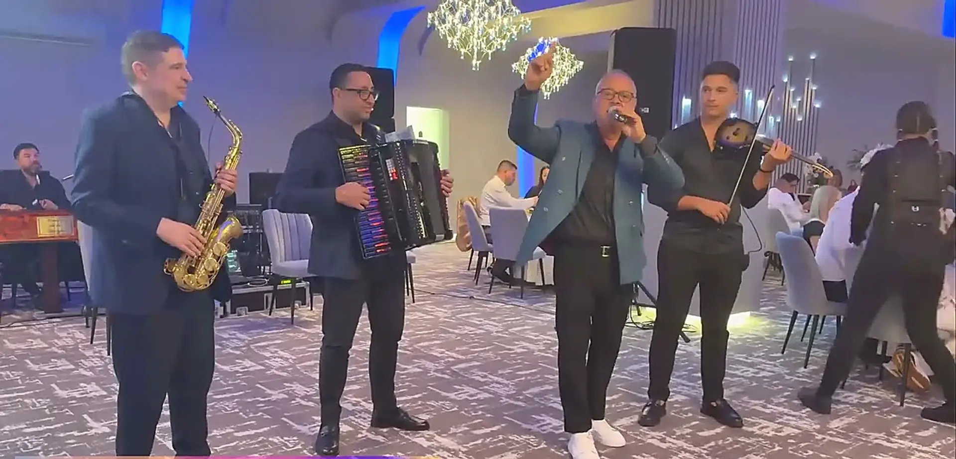 Formația cântând live într-o sală de evenimente, cu saxofon, acordeon și vioară